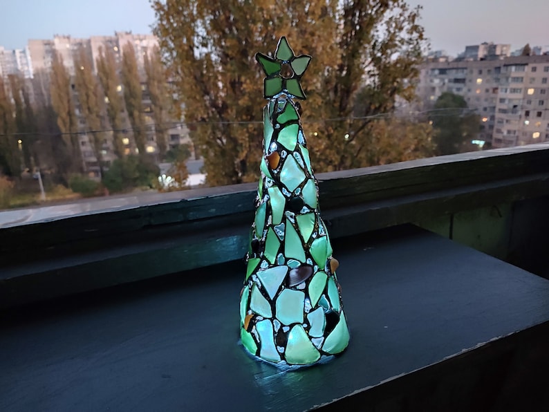 Pode incluir: Uma &aacute;rvore de Natal decorativa feita de pe&ccedil;as de vidro verde e transparente. A &aacute;rvore &eacute; encimada por uma estrela verde e fica sobre uma superf&iacute;cie escura. A &aacute;rvore &eacute; iluminada, criando um efeito brilhante. O fundo mostra uma paisagem urbana.