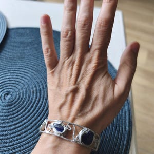 May include: A silver-toned bracelet with three dark blue, irregularly shaped stones. The bracelet has an openwork design with decorative swirls and curves. The bracelet is worn on a wrist.