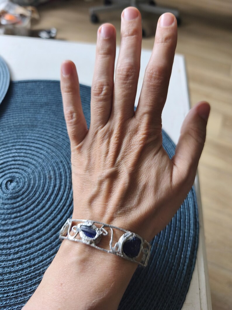 May include: A silver-toned bracelet with three dark blue, irregularly shaped stones. The bracelet has an openwork design with decorative swirls and curves. The bracelet is worn on a wrist.