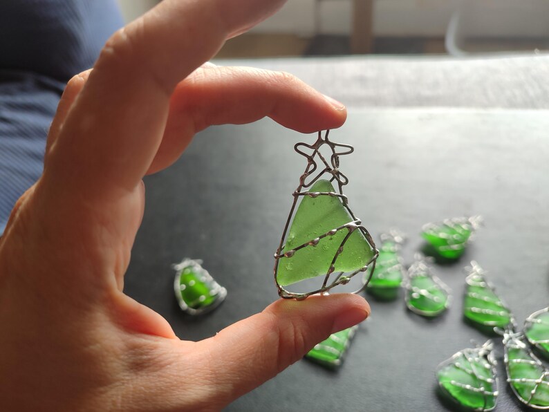 May include: A collection of green glass Christmas tree ornaments wrapped in silver wire. The ornaments are handmade and feature a variety of shapes and sizes.