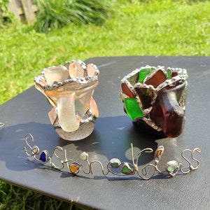 May include: Two small glass candle holders made with stained glass. The holders are in the shape of a vase and have a silver metal rim. The holders are sitting on a black surface with the words "Sea Stained Glass" written in wire with small pieces of sea glass.