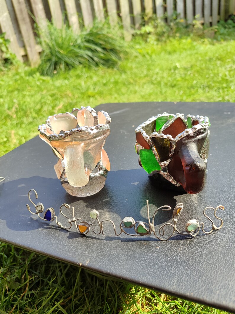 May include: Two small glass candle holders made with stained glass. The holders are in the shape of a vase and have a silver metal rim. The holders are sitting on a black surface with the words "Sea Stained Glass" written in wire with small pieces of sea glass.