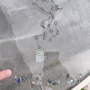 May include: A silver-toned necklace with a cat pendant and a separate wire-formed word, "Sea Stones", with colorful glass stones. The cat pendant is light green with a silver outline. The necklace is on a glass surface.