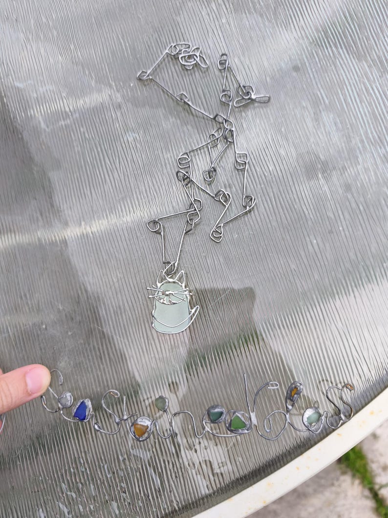 May include: A silver-toned necklace with a cat pendant and a separate wire-formed word, "Sea Stones", with colorful glass stones. The cat pendant is light green with a silver outline. The necklace is on a glass surface.