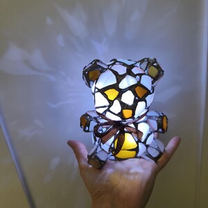 May include: A stained glass teddy bear lamp with a brown ribbon. The lamp is made of white, yellow, and brown glass pieces. The lamp is lit from within, casting a warm glow.