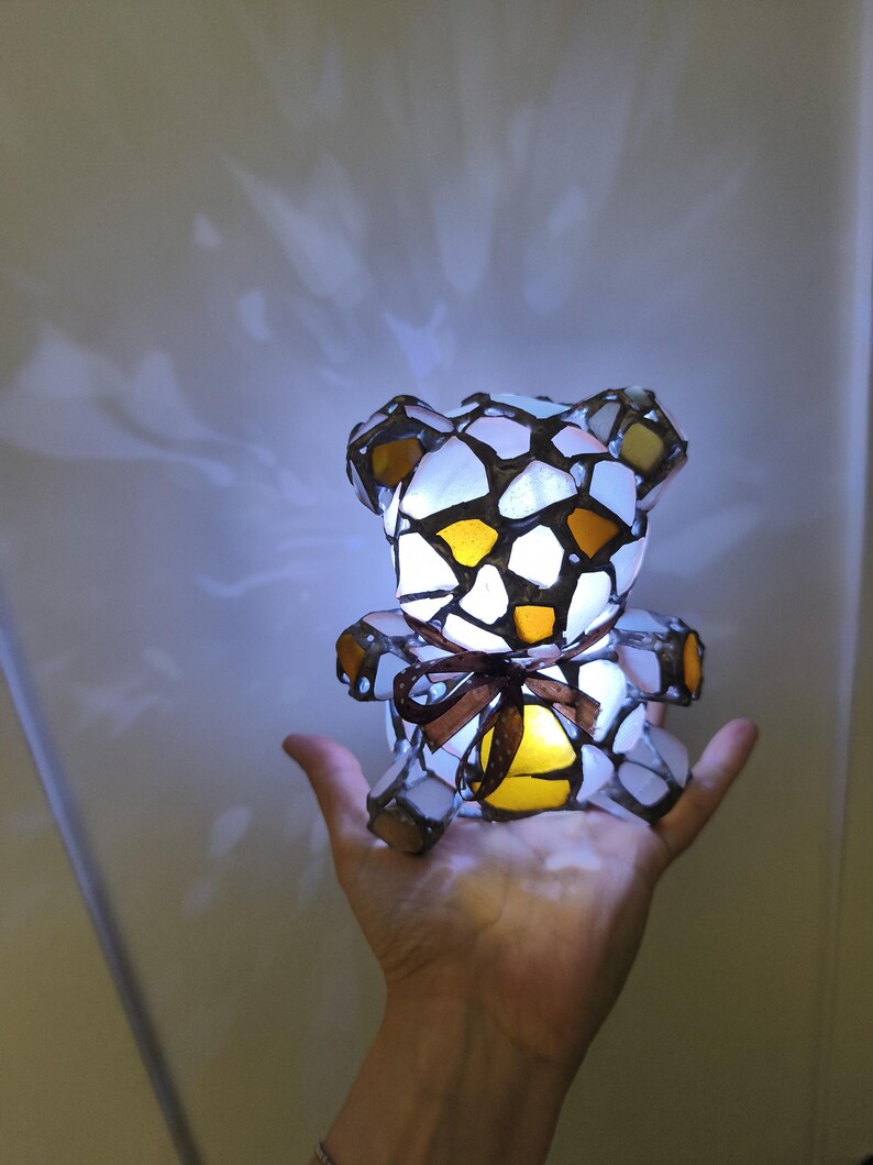 May include: A stained glass teddy bear lamp with a brown ribbon. The lamp is made of white, yellow, and brown glass pieces. The lamp is lit from within, casting a warm glow.