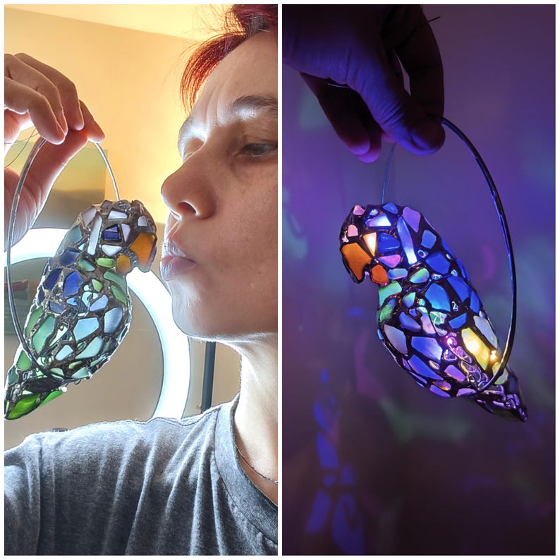 May include: A stained glass parrot ornament with a silver wire loop for hanging. The parrot features a mosaic design with blue, green, and clear glass pieces. The beak is orange. The ornament is held up by a person.