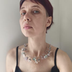 May include: A silver wire necklace featuring a series of irregularly shaped, translucent, and pastel-colored stones. The stones are primarily pink, green, and clear, and are strung along the wire in a delicate arrangement. The necklace is worn against a black tank top.