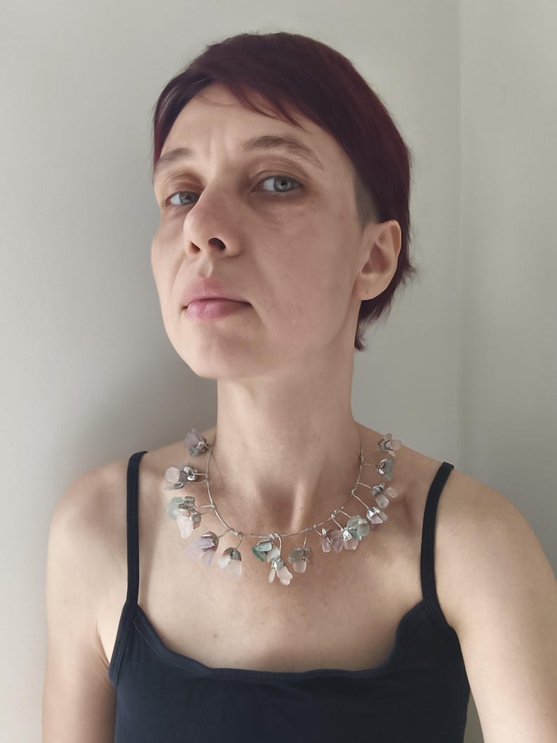 May include: A silver wire necklace featuring a series of irregularly shaped, translucent, and pastel-colored stones. The stones are primarily pink, green, and clear, and are strung along the wire in a delicate arrangement. The necklace is worn against a black tank top.