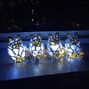 May include: Four stained glass cat figurines with a white, yellow, and blue mosaic design. The figurines are illuminated from within, casting a warm glow.