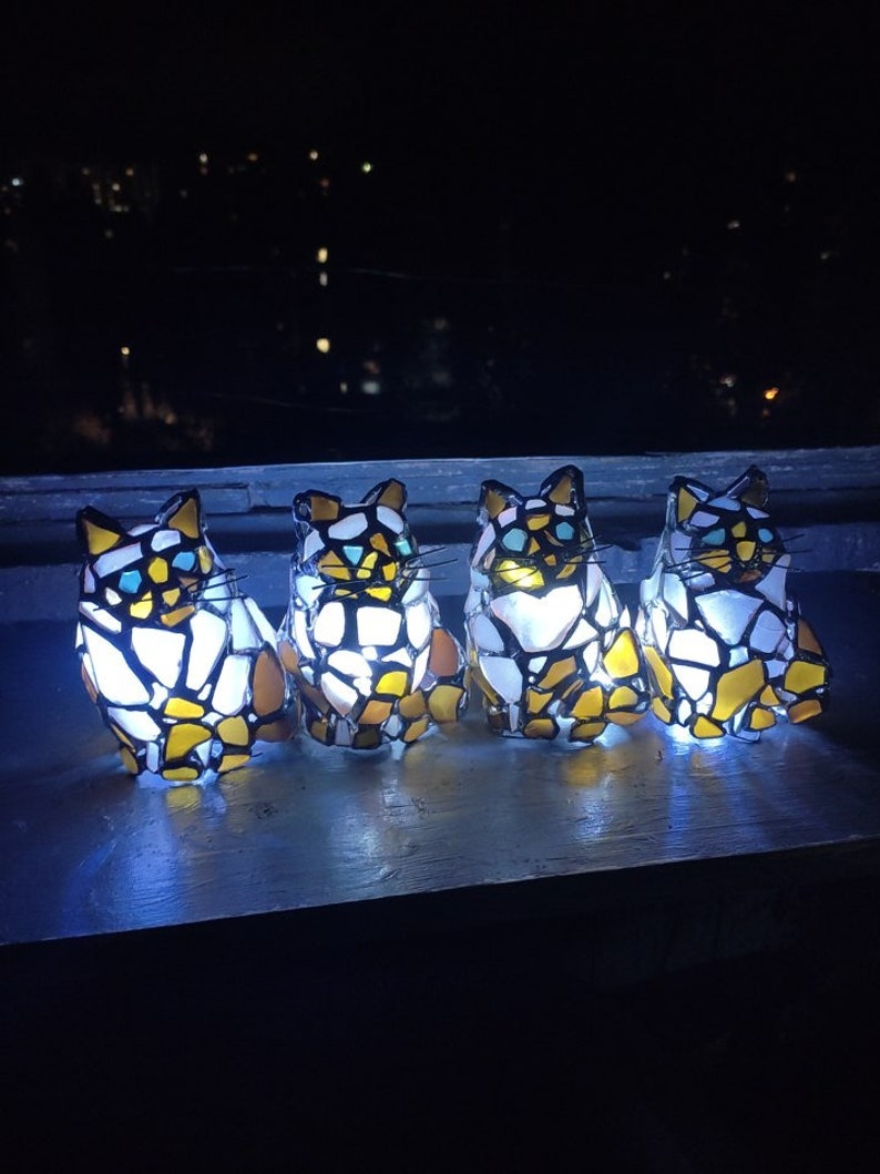 May include: Four stained glass cat figurines with a white, yellow, and blue mosaic design. The figurines are illuminated from within, casting a warm glow.