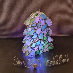 K&ouml;nnte beinhalten: Ein kleiner, beleuchteter Weihnachtsbaum-Anh&auml;nger aus Meerglas und Draht. Der Anh&auml;nger ist in Form eines traditionellen Weihnachtsbaums gestaltet und mit verschiedenen Farben von Meerglas verziert. Der Anh&auml;nger wird von innen beleuchtet und erzeugt so ein warmes Leuchten. Das Wort "Seastone" ist in Draht unter dem Anh&auml;nger geschrieben.