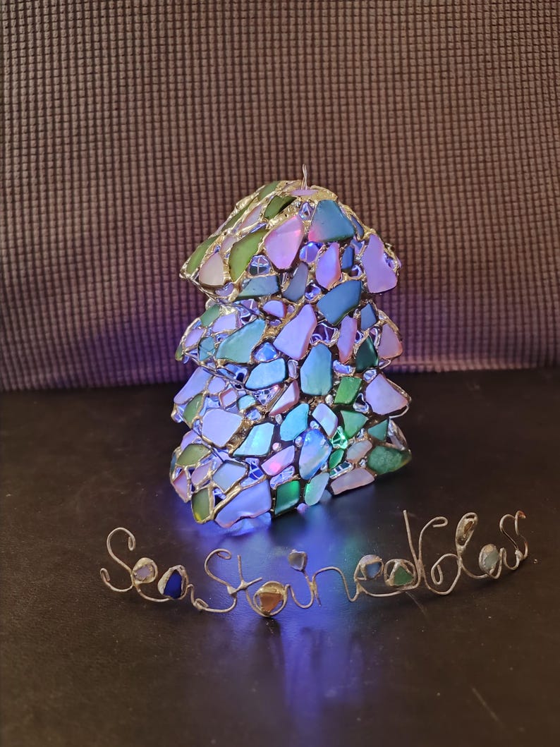 K&ouml;nnte beinhalten: Ein kleiner, beleuchteter Weihnachtsbaum-Anh&auml;nger aus Meerglas und Draht. Der Anh&auml;nger ist in Form eines traditionellen Weihnachtsbaums gestaltet und mit verschiedenen Farben von Meerglas verziert. Der Anh&auml;nger wird von innen beleuchtet und erzeugt so ein warmes Leuchten. Das Wort "Seastone" ist in Draht unter dem Anh&auml;nger geschrieben.