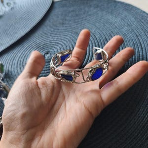 May include: A silver-toned cuff bracelet with irregular edges and embedded blue glass stones. The bracelet is open at the bottom and is held in a hand against a blue background. The bracelet's design is intricate and eye-catching.