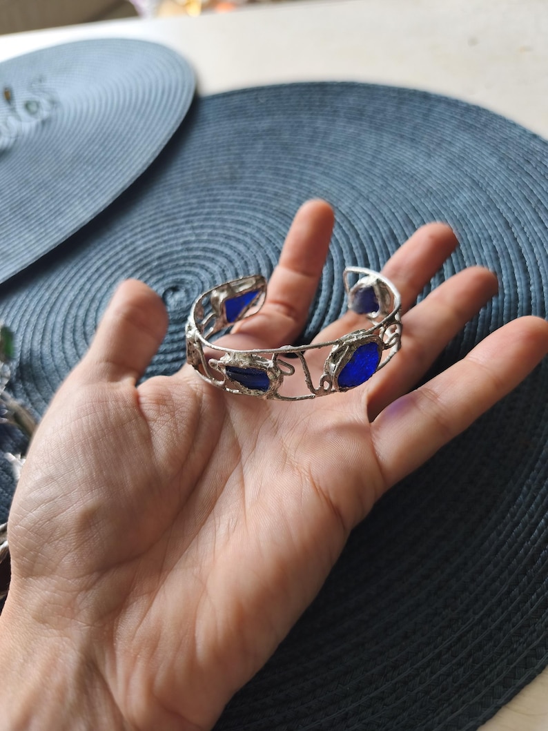 May include: A silver-toned cuff bracelet with irregular edges and embedded blue glass stones. The bracelet is open at the bottom and is held in a hand against a blue background. The bracelet's design is intricate and eye-catching.