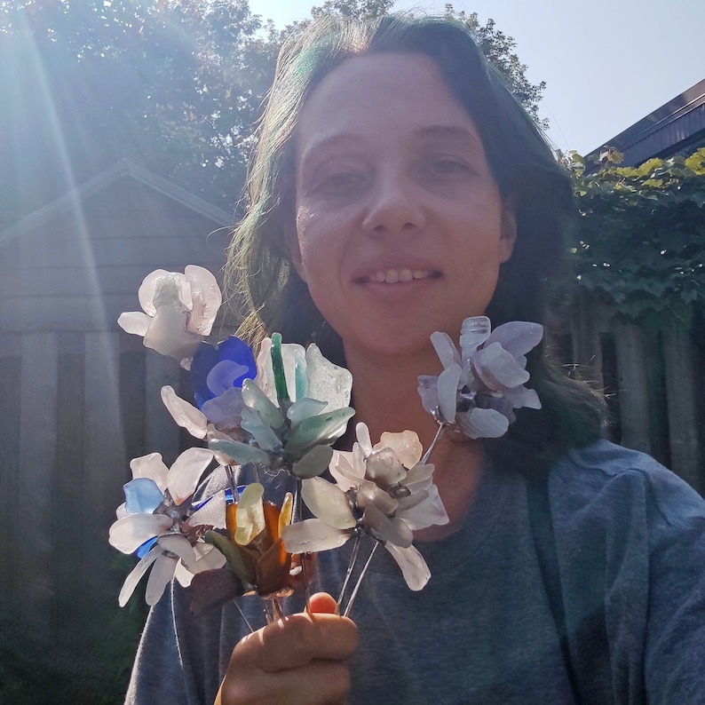 Op de afbeelding: Een boeket handgemaakte bloemen gemaakt van zeeglas. De bloemen zijn in tinten wit, blauw en groen. De bloemen worden vastgehouden door een persoon met groen haar.