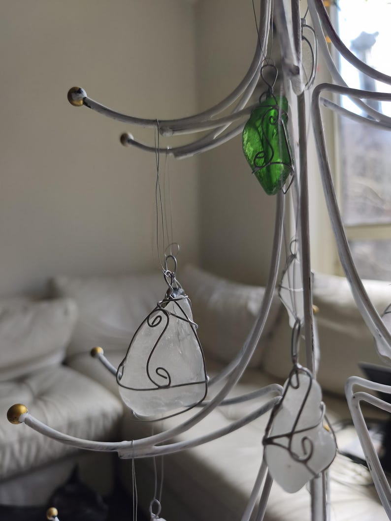May include: A white metal tree-shaped display stand with ornaments. The ornaments are made of clear and green glass, wrapped in wire. The stand has gold-colored tips on the branches. The ornaments are teardrop-shaped.