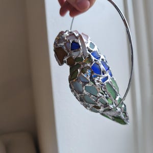 May include: A colorful, stained glass parrot sculpture with a silver metal frame. The parrot is composed of various shades of blue, green, brown, and white glass pieces. The sculpture is suspended by a silver metal ring.