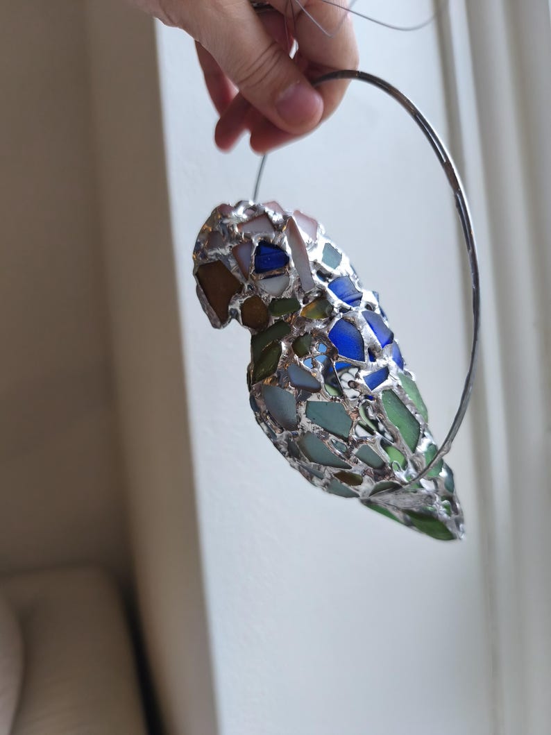 May include: A colorful, stained glass parrot sculpture with a silver metal frame. The parrot is composed of various shades of blue, green, brown, and white glass pieces. The sculpture is suspended by a silver metal ring.