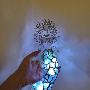 Puede incluir: Una figura de sirena hecha de alambre y vidrio marino iluminado. La figura presenta una cabeza y cabello esculpidos en alambre, un corpi&ntilde;o de vidrio marino y una cola construida con m&uacute;ltiples piezas de vidrio azul claro unidas con soldadura.