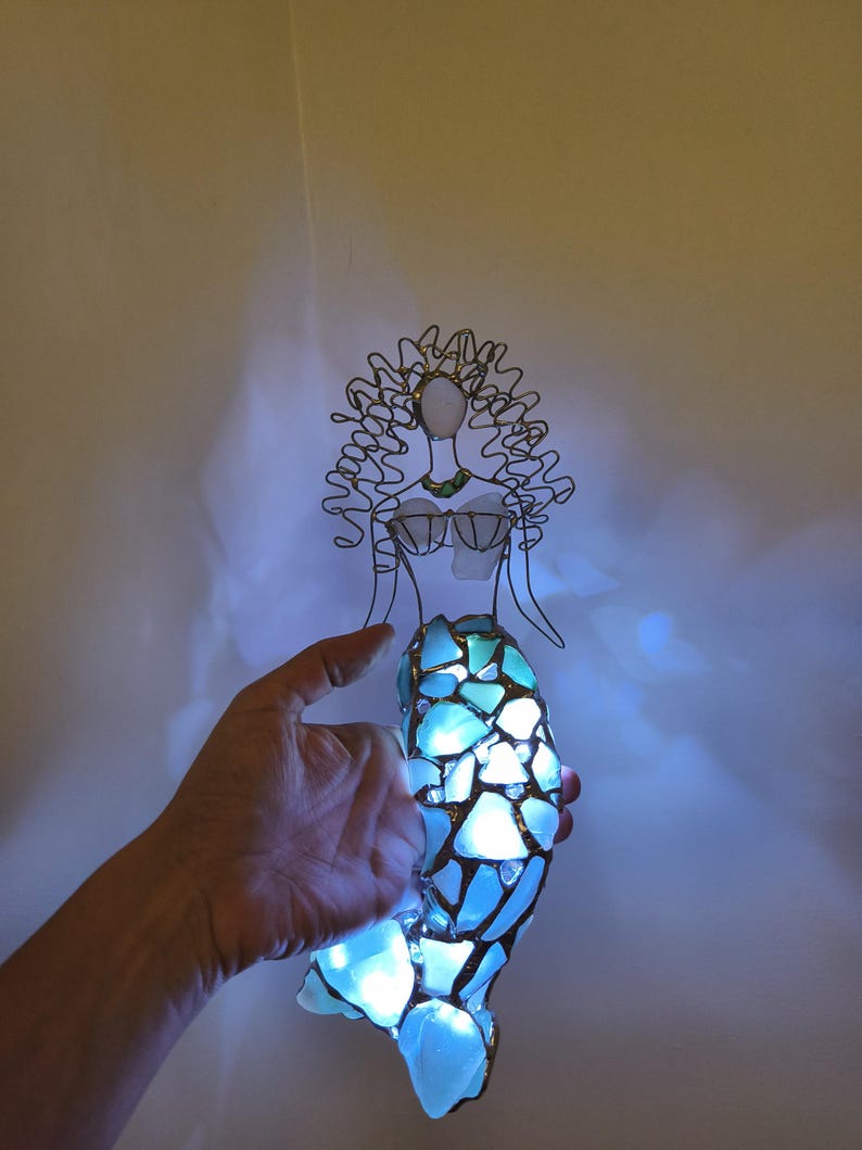 Puede incluir: Una figura de sirena hecha de alambre y vidrio marino iluminado. La figura presenta una cabeza y cabello esculpidos en alambre, un corpi&ntilde;o de vidrio marino y una cola construida con m&uacute;ltiples piezas de vidrio azul claro unidas con soldadura.