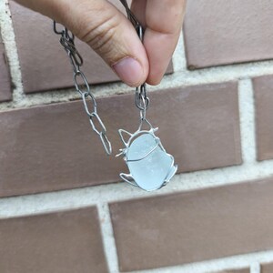 May include: A handmade necklace featuring a pendant made of sea glass encased in silver-colored wire. The pendant is a light blue color and is suspended from a silver-colored chain.