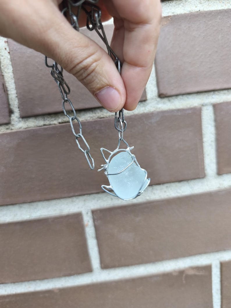 May include: A handmade necklace featuring a pendant made of sea glass encased in silver-colored wire. The pendant is a light blue color and is suspended from a silver-colored chain.