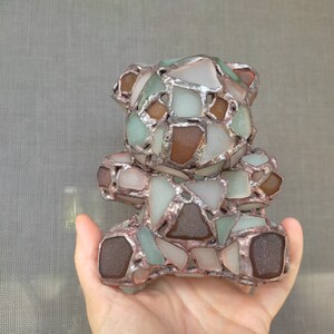 May include: A handcrafted teddy bear sculpture made from sea glass pieces in shades of brown, green, and white, held in a hand. The sea glass is set in a silver-colored metal frame, creating a mosaic effect. The bear is a unique decorative item.