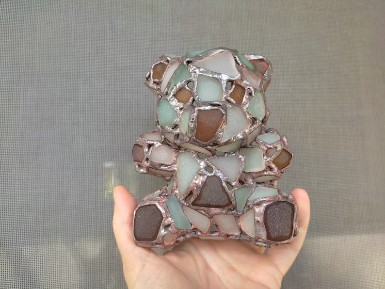 May include: A handcrafted teddy bear sculpture made from sea glass pieces in shades of brown, green, and white, held in a hand. The sea glass is set in a silver-colored metal frame, creating a mosaic effect. The bear is a unique decorative item.