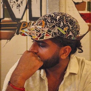 May include: A mosaic-style baseball cap with a black base and colorful glass pieces. The cap features a curved brim and a decorative pattern. The background includes graffiti art and a person wearing a light-colored shirt.