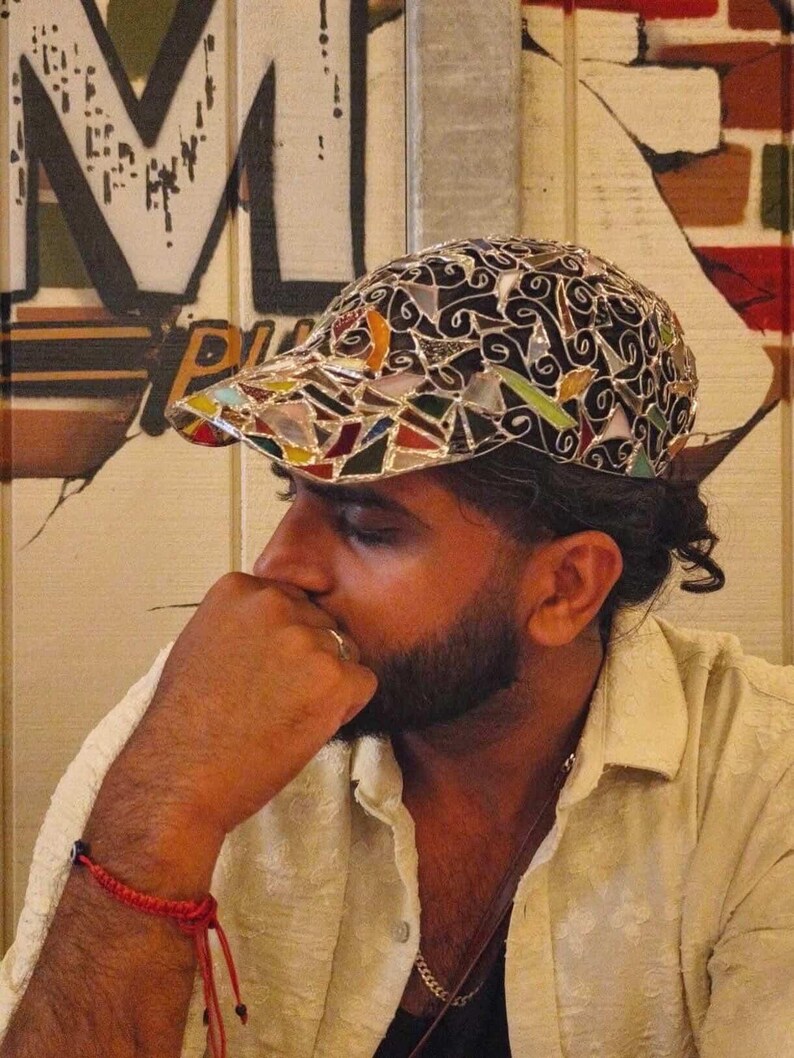 May include: A mosaic-style baseball cap with a black base and colorful glass pieces. The cap features a curved brim and a decorative pattern. The background includes graffiti art and a person wearing a light-colored shirt.
