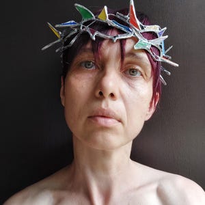 May include: A crown made of stained glass pieces in red, yellow, blue, and green, held together by silver-toned metal solder. The crown features sharp, pointed edges and a jagged, irregular structure that rests atop the head.