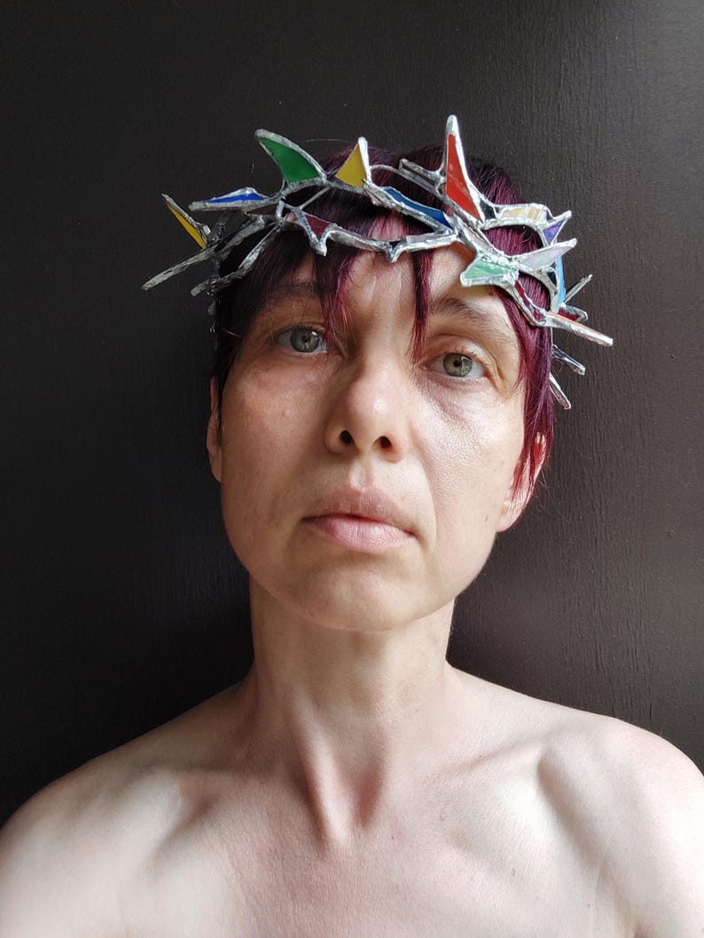 May include: A crown made of stained glass pieces in red, yellow, blue, and green, held together by silver-toned metal solder. The crown features sharp, pointed edges and a jagged, irregular structure that rests atop the head.