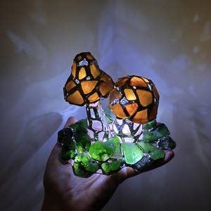 May include: A decorative mushroom lamp with two illuminated caps. The caps are made of amber-colored glass pieces, outlined in black, and sit atop a base of green glass. The lamp is held in a hand, with a soft glow emanating from within.