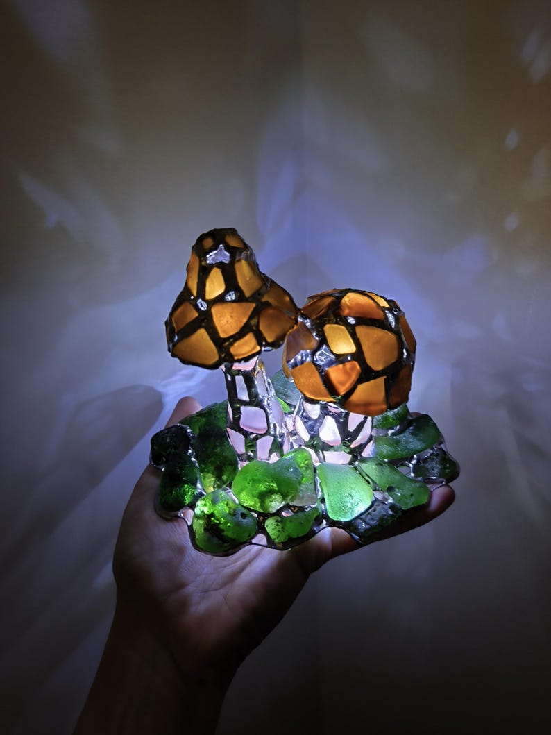 May include: A decorative mushroom lamp with two illuminated caps. The caps are made of amber-colored glass pieces, outlined in black, and sit atop a base of green glass. The lamp is held in a hand, with a soft glow emanating from within.