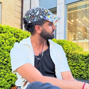 May include: A patterned cap with a black and white base and colorful geometric shapes. The cap is worn with a white shirt over a black top. A brown leather bag is visible.