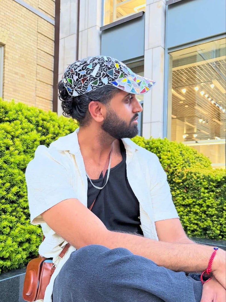 May include: A patterned cap with a black and white base and colorful geometric shapes. The cap is worn with a white shirt over a black top. A brown leather bag is visible.