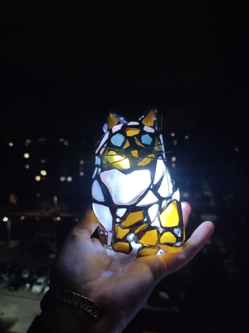 May include: A stained glass cat figurine with yellow, white, and blue glass pieces. The cat is illuminated from within, casting a warm glow.