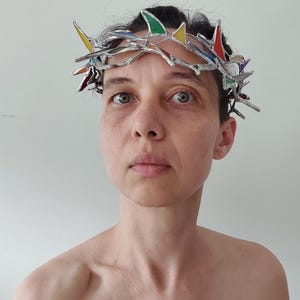 May include: A crown-shaped headpiece constructed from silver-toned metal and small, multi-colored stained glass shards in red, yellow, green, and blue. The structure features sharp, pointed segments arranged in a circular, thorn-like pattern.