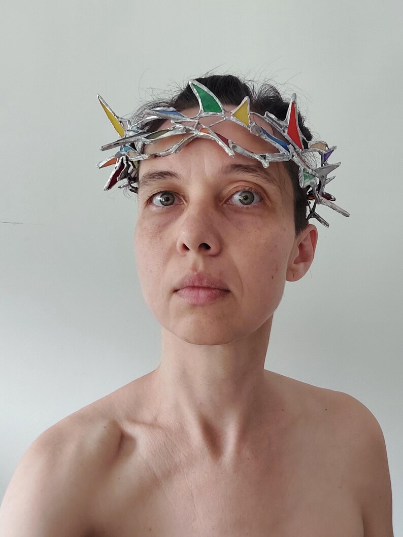 May include: A crown-shaped headpiece constructed from silver-toned metal and small, multi-colored stained glass shards in red, yellow, green, and blue. The structure features sharp, pointed segments arranged in a circular, thorn-like pattern.