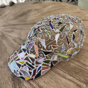 May include: A mosaic baseball cap with a curved brim. The cap is made of small, irregularly shaped pieces of colored glass, including red, blue, green, and yellow. The pieces are set in a silver-colored metal frame.