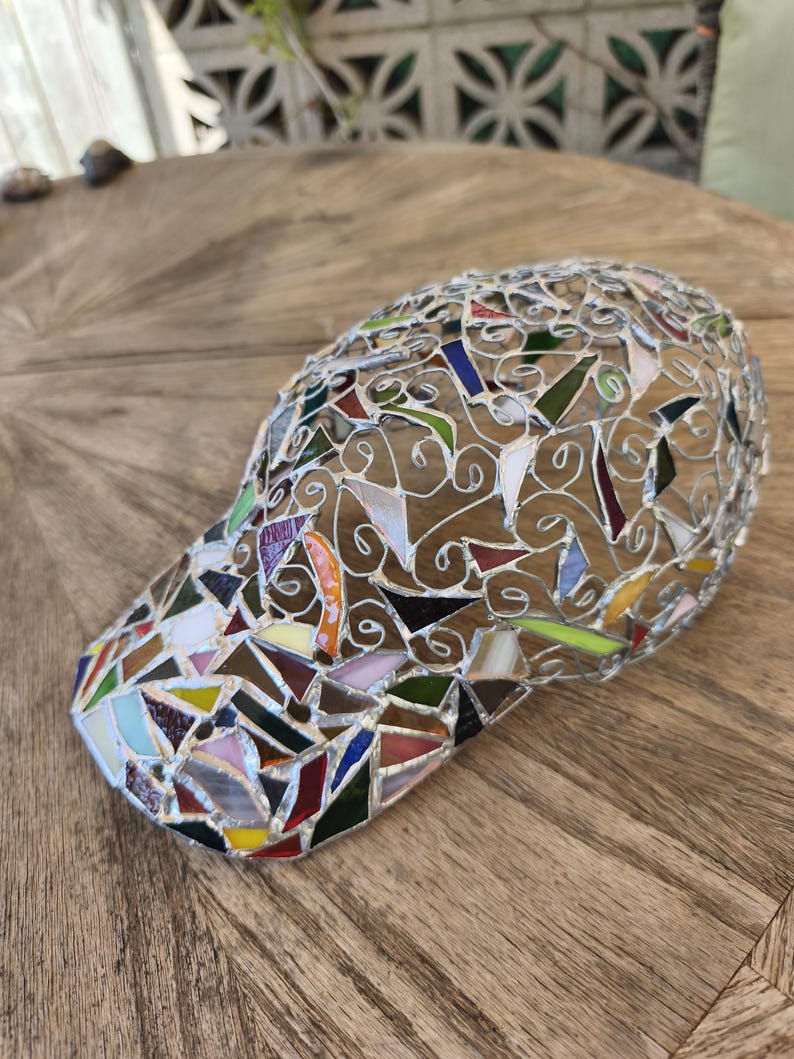 May include: A mosaic baseball cap with a curved brim. The cap is made of small, irregularly shaped pieces of colored glass, including red, blue, green, and yellow. The pieces are set in a silver-colored metal frame.