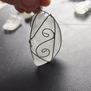 May include: A clear, irregularly shaped pendant made of sea glass, encased in a decorative wire frame. The wire has a swirling design on the front. Other similar pendants are visible in the background. The pendant is held by a hand.