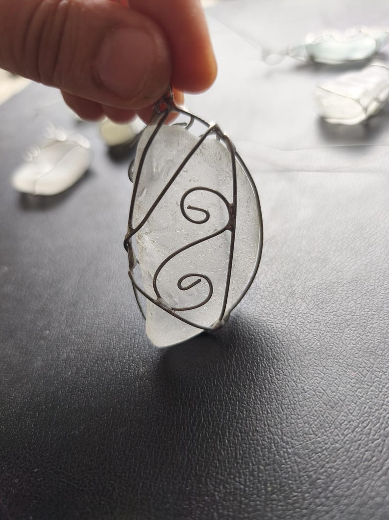 May include: A clear, irregularly shaped pendant made of sea glass, encased in a decorative wire frame. The wire has a swirling design on the front. Other similar pendants are visible in the background. The pendant is held by a hand.