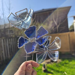 May include: A stained glass flower sculpture with blue and clear petals. The glass is held together with silver-coloured wire, and the sculpture is mounted on a metal stem. The flowers are displayed outdoors in front of a green lawn.