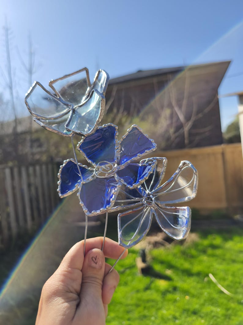 May include: A stained glass flower sculpture with blue and clear petals. The glass is held together with silver-coloured wire, and the sculpture is mounted on a metal stem. The flowers are displayed outdoors in front of a green lawn.
