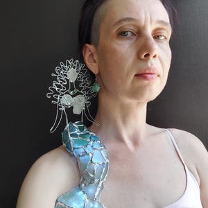 Puede incluir: Una figura decorativa de sirena hecha de alambre plateado y trozos de vidrio marino en tonos azules y verdes. La sirena presenta una cabeza y un torso formados por alambre con una cola de mosaico hecha de piezas de vidrio transl&uacute;cido.