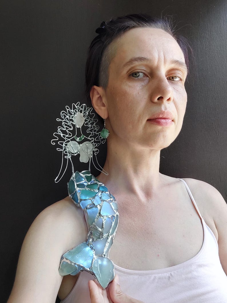 Puede incluir: Una figura decorativa de sirena hecha de alambre plateado y trozos de vidrio marino en tonos azules y verdes. La sirena presenta una cabeza y un torso formados por alambre con una cola de mosaico hecha de piezas de vidrio transl&uacute;cido.