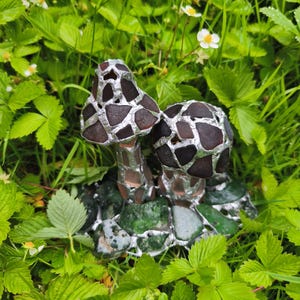 May include: A pair of decorative mushroom sculptures with brown and silver mosaic caps and stems. The mushrooms are set on a base of green stones and are surrounded by green foliage and small white flowers.