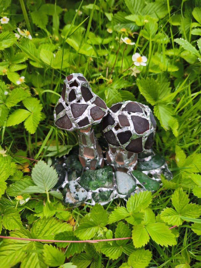 May include: A pair of decorative mushroom sculptures with brown and silver mosaic caps and stems. The mushrooms are set on a base of green stones and are surrounded by green foliage and small white flowers.